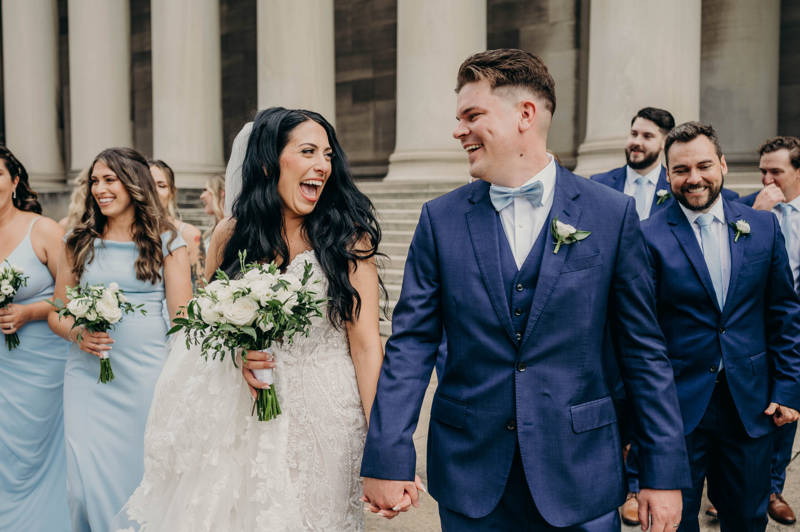 Bride and groom laughing and holding hands with their wedding party in Pittsburgh, PA. The bride wears a lace gown and holds a white floral bouquet, while the groom wears a navy blue suit with a bow tie. Captured by CorianneCPhotography, a Pittsburgh wedding photographer.